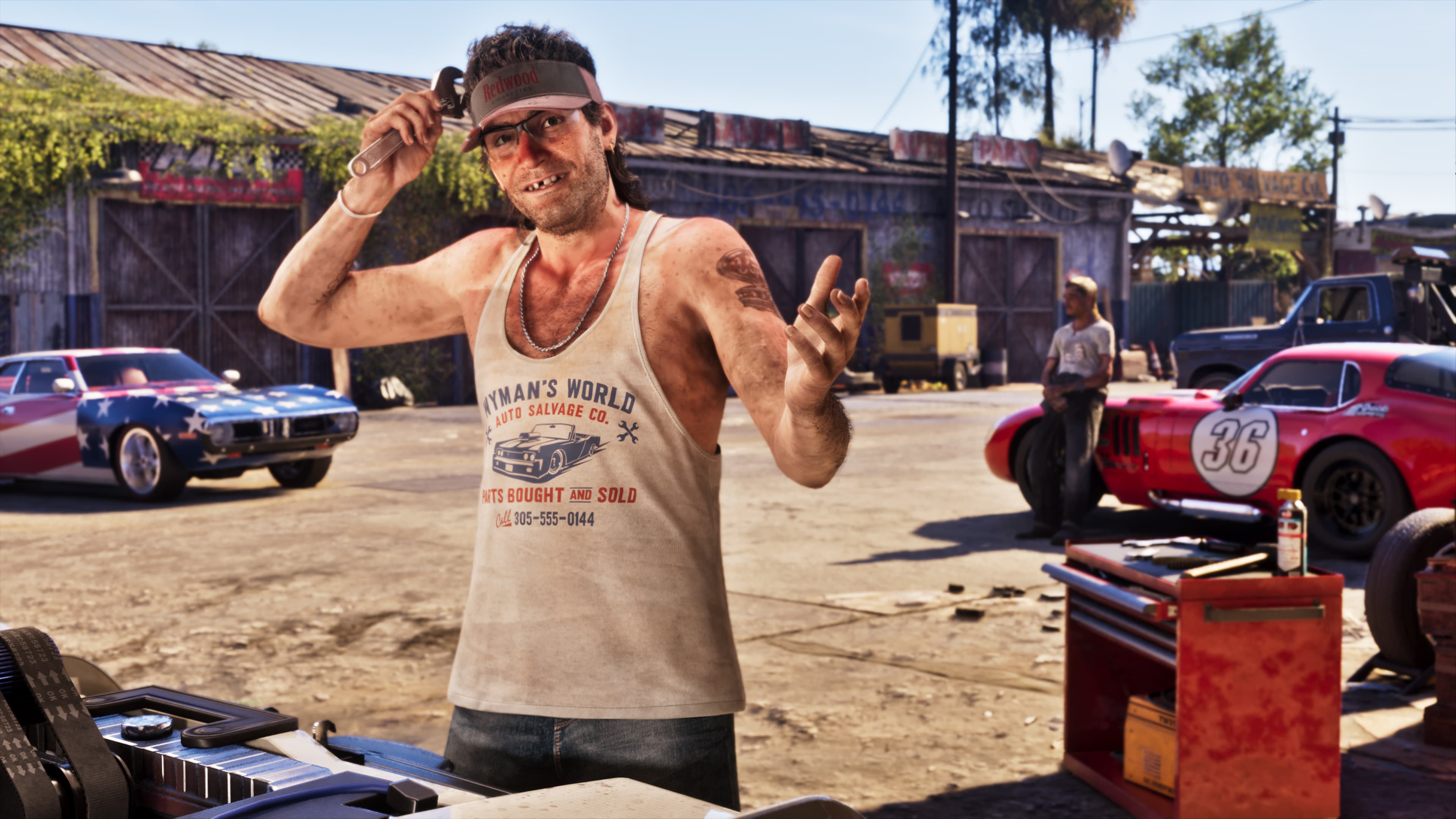 A man in a vest holding a phone in a dirt yard surrounded by classic cars